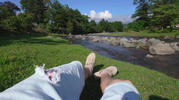 年轻英俊的男子在湖边或河边的空地上休息 第一人称在阳光下享受大自然 双腿交叉的男人躺在美丽的河岸上 — 图库视频影像