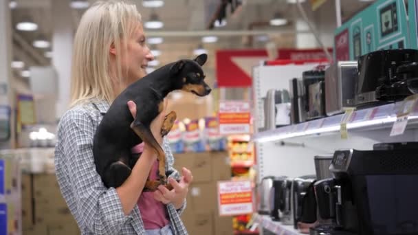 年轻漂亮的金发姑娘带着她的小宠物狗在超市或商店里走来走去 拿起家用电器 孩子的自由 无子女 — 图库视频影像