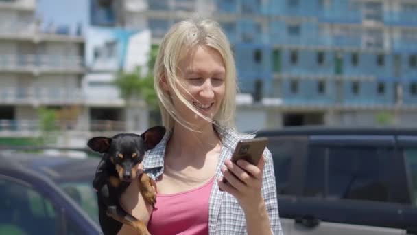 与宠物狗在一起的年轻女人的特写镜头 拿着她的手机 看着屏幕 千禧年数位生活方式的概念 他们在阳光明媚的日子漫步在城市的街道上 无子女 — 图库视频影像