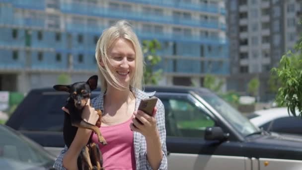 与宠物狗在一起的年轻女人的特写镜头 拿着她的手机 看着屏幕 千禧年数位生活方式的概念 他们在阳光明媚的日子漫步在城市的街道上 无子女 — 图库视频影像