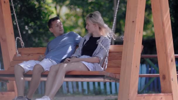 一对年轻夫妇骑在室外公园的木制秋千上 男人抚摸女孩的头发 恋爱中的已婚夫妇很享受 年轻的家庭和情人 — 图库视频影像