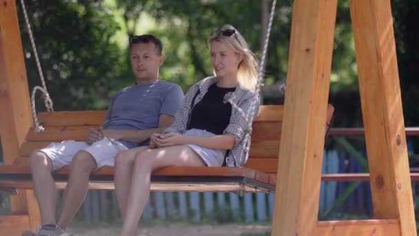 屋外公園で木製のスイングに乗っている若いカップル男性と女性 ストローク ガールズの髪 恋に浮かぶ夫婦は楽しんでいる 若い家族と恋人 — ストック動画