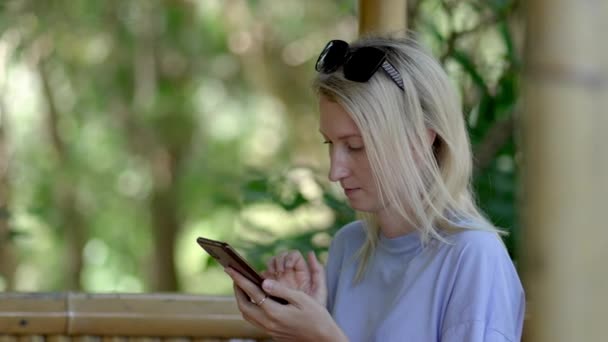 若い美しい金髪の女性は ガゼボで公園に座って 携帯電話 スマートフォンを使用してテキスト 晴れた夏の日 彼女は新鮮な空気の中で働いたりリラックスしたりします 学生について — ストック動画