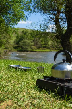 Alüminyum plakalar, kupalar, yeşil çimlerin üzerinde çaydanlık tezgahı olan bir kamp gaz ocağı, bir dağ nehrinin arka planına ve tuzlu bir günde ağaçlara yakın çekim. Arabayla seyahat ederken yemek yapmak..