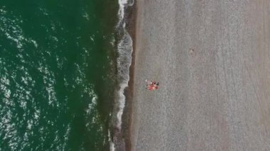 Çakıl taşlı plajdaki birkaç insanın hava görüntüsü. Boş Macera. Drone deniz dalgaları vuracak ve balaylarında aşık çiftlerin etrafında uçacak. Meet or Lovers. Okyanusta rahatla. Yeşil deniz ve taşlar.