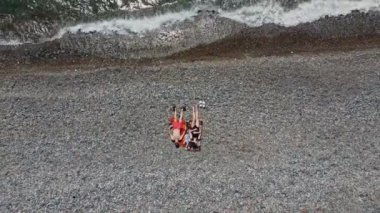 Çakıl taşlı plajdaki birkaç insanın hava görüntüsü. Boş Macera. Drone deniz dalgaları vuracak ve balaylarında aşık çiftlerin etrafında uçacak. Meet or Lovers. Okyanusta rahatla. Yeşil deniz ve taşlar.