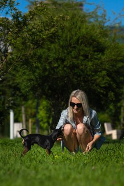 Siyah oyuncak teriyeri genç sarışın bir hostese sarı bir top getirdi. Kadın topu köpeğin getirmesi gerçeğine lezzet katıyor. Evcil bir hayvanla açık hava oyunları. Dikey fotoğraf