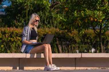 Şortlu, tişörtlü, güneş gözlüklü güzel sarışın bir kadın güneşli bir yaz gününde bir parkta dizüstü bilgisayarda çalışıyor. Öğrenci ya da serbest çalışan biri şehir parkında dizüstü bilgisayarda çalışıyor.