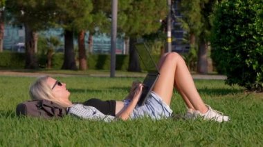 Güzel bir öğrenci ya da serbest çalışan çimlerin üzerinde yatarken dizüstü bilgisayar kullanıyor. Eğitim kavramı. Park işkadını ya da çalışanı çimenlerin üzerinde uzanan gülümseyen genç bir kızın portresi..