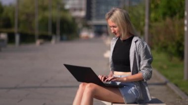 Serbest çalışan kadın ya da iş kadını parkta bankta otururken dizüstü bilgisayar kullanır. İnternette çalışan ya da bilgisayarla çalışıp öğrenen genç bir kadın. Güneşli yaz günü.