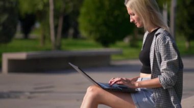 Çimlerde oturan bir kadın, dizüstü bilgisayar ve üniversite öğrencisi eğitim materyalleri ve eğitim okuyor. Dışarıda çalışan ve bilgisayar kullanan mutlu bir kadın. Parktaki bankta oturmuş bilgisayar kullanıyor.