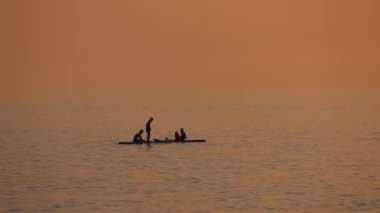 Bir grup insan gün batımında SUP 'ye biner, tekneler yüzer. Resimli turuncu gün batımı güneşi yansıtıyor. Adaların tarihöncesi. Özgürlük, mutluluk. Seyahat, Egzotik Turizm.