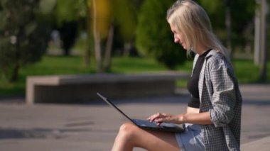 Serbest çalışan kadın ya da iş kadını parkta bankta otururken dizüstü bilgisayar kullanır. İnternette çalışan ya da bilgisayarla çalışıp öğrenen genç bir kadın. Güneşli yaz günü.