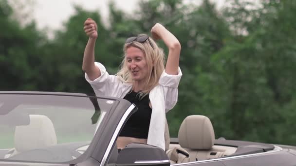 手を挙げてコンバーチブルで踊る女性 夏の旅行で風の強い日を楽しんでいる若い女性のハンドヘルド映像 彼女は楽しい旅行をしている — ストック動画