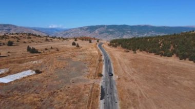 Drone, dağ geçidine kırsal yol boyunca giden ve Türkiye 'deki boş tarlalarda giden otomobili izliyor. Dağların eteğindeki tarım arazisi. Sıcak güneşli bir gün, romantizm tatilde arabayla seyahat etmek.