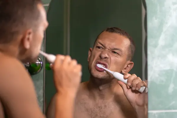 Young man with a bare torso and hairy chest brushes his teeth with an electric brush while looking in the bathroom mirror. Morning rituals, personal hygiene