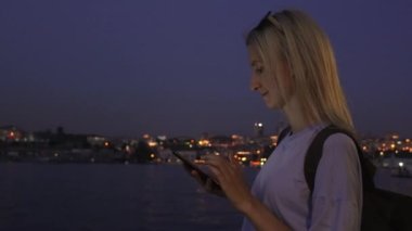 Şehir sokağında duran akıllı telefona bakan genç ve güzel bir kadın. Gece lambalarının arka planında cep telefonu kullanan kadın portresi. Beyaz tenli sarışın akıllı telefon ekranına bakıyor..