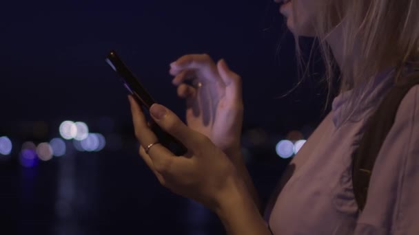 夜の街路に立っているスマートフォンを見ている美しい若い女性 夜の街灯の背景に携帯電話を使用する女性の肖像画 スマートフォン画面を見ている白人ブロンド — ストック動画
