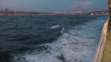 Güverte ve ahşap teknenin üst manzarası geceleri denize bakan şehirde seyrediyor. Gemi köpüklü beyaz deniz dalgalarını kesiyor. Beyaz köpüklü hareket eden pervane motoru ile beyaz kabarcık fırladı
