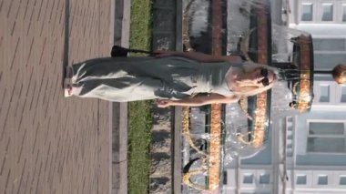 Şık bir iş elbisesi içinde, fıskiyenin arka planında yeşil parkta yürüyen sarışın bir kadının tam boy portresi. Şehir caddesinde yürüyen mutlu genç bir kadın. Dikey video