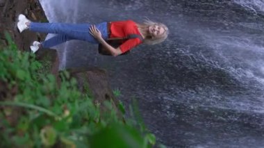 Şelalenin önünde duran kadının arka görüntüsü. Sırt çantalı kadın dağ nehri şelalesinin yanında duruyor ve doğanın gücünün dalgalanmasının tadını çıkarıyor. Seyahat, Seyahat ve Doğanın Kavramsal Görüntüsü.