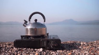 Portatif gaz sobasında kaynayan metal çaydanlığın manzarası, sakin gölün arka planına ve şafak vakti dağlara karşı, doğada yolculuk ve dinlenme..