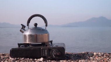 Portatif gaz sobasında kaynayan metal çaydanlığın manzarası, sakin gölün arka planına ve şafakta dağlara karşı, doğada yolculuk ve dinlenme. Dikey video