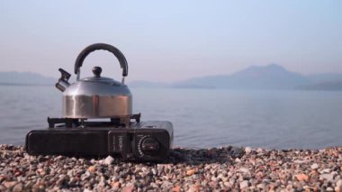 Portatif gaz sobasında kaynayan metal çaydanlığın manzarası, sakin gölün arka planına ve şafakta dağlara karşı, doğada yolculuk ve dinlenme. Dikey video