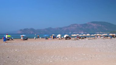 IZMIR, TURKEY - 1 Eylül 2023: Patara plajında plaj gününün tadını çıkaran, güneşlenen ve deniz kenarında dinlenen bir grup insan güneşli bir günde resimli dağ manzarasına hayran kaldılar.