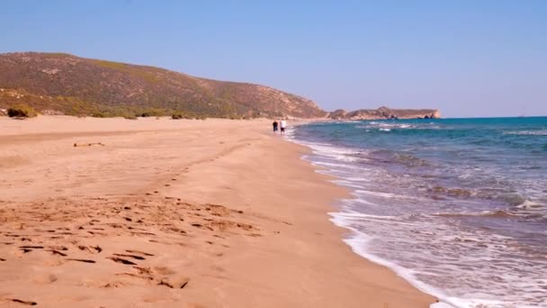 海の波と山々を背景にした砂浜の景色は 素晴らしい自然景観を生み出しています 晴れた日 見知らぬ二人が海岸を歩いている 人なしで空 — ストック動画