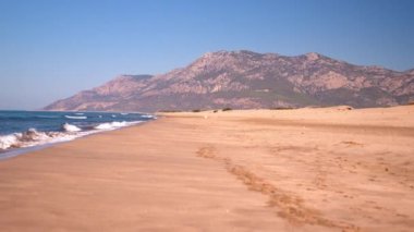 Temiz deniz suyuyla altın kumsal manzarası ve arka planda görkemli dağlar, çarpıcı bir doğal manzara yaratıyor. Güneşli bir gün ve dalgalar. Boşluğu kopyala.