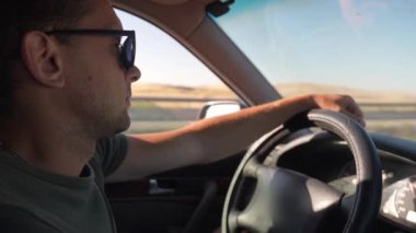 Kabinin içinde araba kullanan adam var. Sürücünün ellerinin direksiyonda yatışı, etraftan geçen çevreye ve manzaraya bakış..