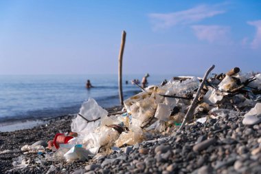 Toplanan bir grup selofan çantanın yakın çekimi, plastikler arka plandaki çakıl taşı plajında insanlar dikkatsiz bir şekilde yüzüyorlar. Ekoloji kavramı, doğayı koruyalım..