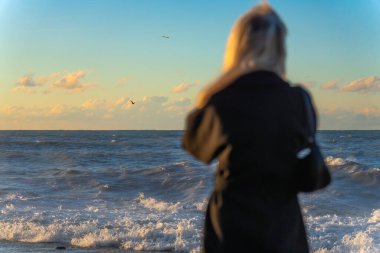 Siyah ceketli sarışın genç bir kadın, gün batımında fırtınalı bir denize ya da okyanusa bakıyor. Seçici bir odak noktası. Denizin ufkuna bakan bir kadının arkasından çekilmiş bir fotoğraf.