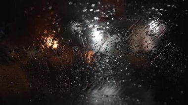 Arabanın ön camına yağmur damlası. Akşam şehrinin caddelerinde duran bir araba, arka planda bulanık trafik ışığı ve sokak lambalı arabaları. Siluet adam ya da hırsız ön kaputun önünde koşuyor..