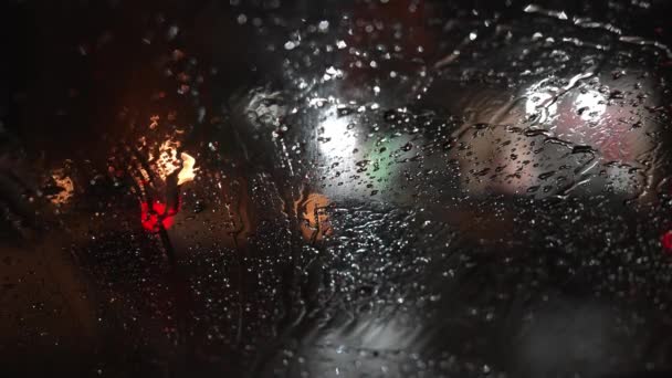 車のフロントガラスに雨が降る 夜の街の通りに立っている車 交通信号のぼやけた背景と街灯で車を移動 — ストック動画