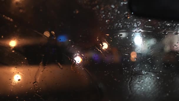 車のフロントガラスに雨が降る 夜の街の通りに立っている車 交通信号のぼやけた背景 街灯で車を動かす 点滅するライトが付いている通る警察か救急車 — ストック動画