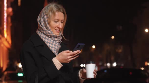 穿着时髦衣服的女人偷东西 一边看智能手机 一边在城市灯光的衬托下 用一次性纸杯盛放热咖啡或茶 一边在室外享受新鲜空气和周末 — 图库视频影像