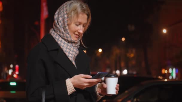 年轻女子一边喝着咖啡一边用她的手机走在城市的街道上 咖啡店和外卖咖啡在城里走来走去 — 图库视频影像