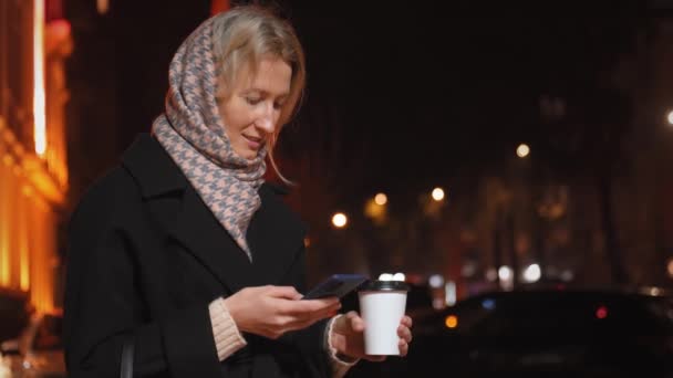 时尚女人在偷东西 沉浸在她的智能手机 享受热饮咖啡或比在城市灯光背景下的一次性纸杯 享受外面的空气和周末 — 图库视频影像