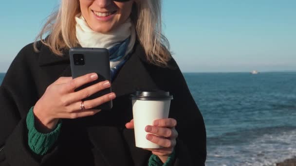 海岸を歩いている間 携帯電話を使ってコーヒーを飲むコートの若い女性 ビジネスマン または学生 コーヒー ビーチ オープンな海岸を歩きます 週末のリラックス テイクアウトコーヒー — ストック動画