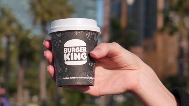 ジョージア州バトゥミ 2024年 バーガーキングのテイクアウトから手札にコーヒーを入れた若い女性 紙コップでキングコーヒーバーガー 女性の手で輝かしい都市の背景 — ストック動画