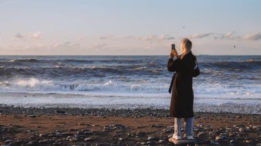 Denizin önünde duran paltolu kadının arka görüntüsü. Cep telefonu kamerasıyla denizi çekiyoruz. Üzgün ve duygusal bir adam mesafeye bakıyor. Gemi ufukta görünüyor..