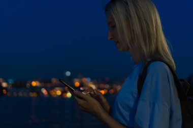 Akıllı telefonlu genç bir kadın, şehir ışıkları ve denizin bulanık arka planında sırt çantası. Cep telefonuyla gülümseyen bir kadının portresi, sosyal ağlara mesajlar yollamak, online alışverişler yapmak.