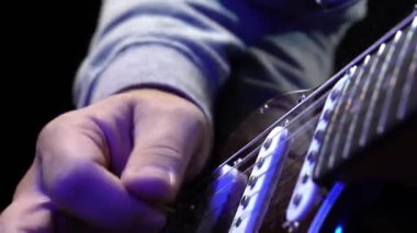 Elektro gitar çalan bir adam, insan ellerine yakın çekim tutkulu bir melodi yaratıyor, parmakları teller ve perdeler üzerinde sorunsuzca hareket ediyor. Neon Mavi Işık.
