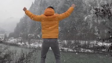 Genç adam kış havasının tadını çıkarıyor dağlarda, nehrin ve ormanın arka planında kollarını açmış, gülümsüyor. Nehir kıyısında bir adam, kar yağıyor.