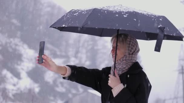 写真を撮ったり 山でセルフィーを撮ったりする若い女性 雪の傘の下で雪の森で携帯電話でビデオ通話をするウィンターウォークガールを楽しんでいます 返された動き — ストック動画