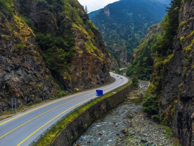 Yüksek kayalık yeşil dağlar ve nehirler arasında yol tabelası olan arabasız güzel asfalt yollar, insansız hava aracı manzarası. Yolun ve dağların güzel manzarası.