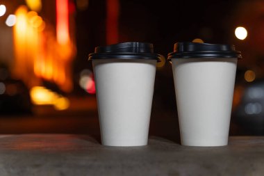 Bulanık bir gece şehrinin arka planında kahve ya da çay eşliğinde beyaz çevre dostu geri dönüşümlü kağıt bardakların yakın çekimi. Kahveyi yanına al..