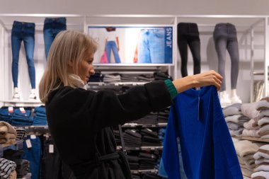 Paltolu güzel, sarışın bir kadın alışveriş merkezinde kıyafet seçiyor.